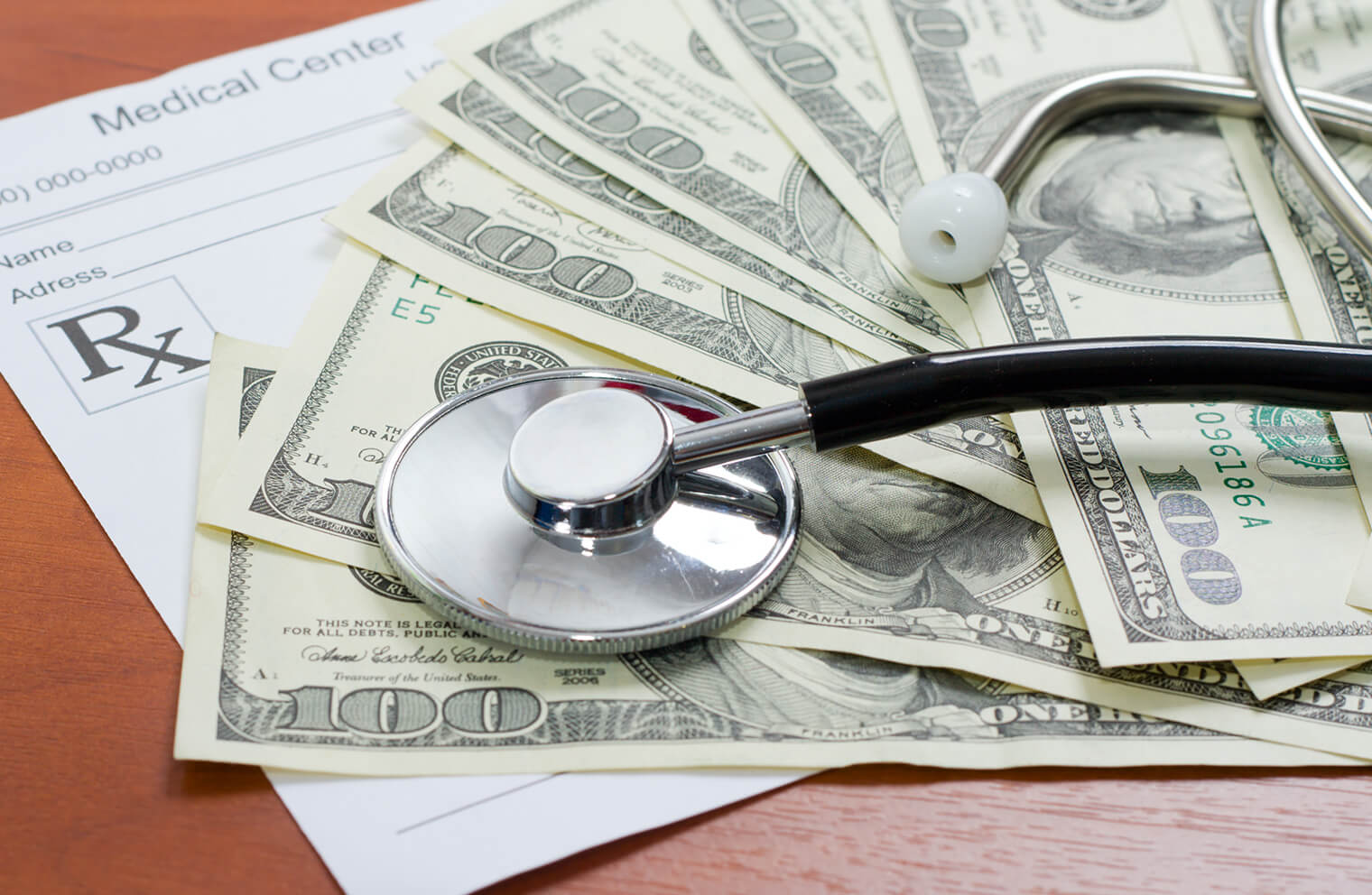 Close up picture of a stethoscope on top of $100 bills and a prescription note.
