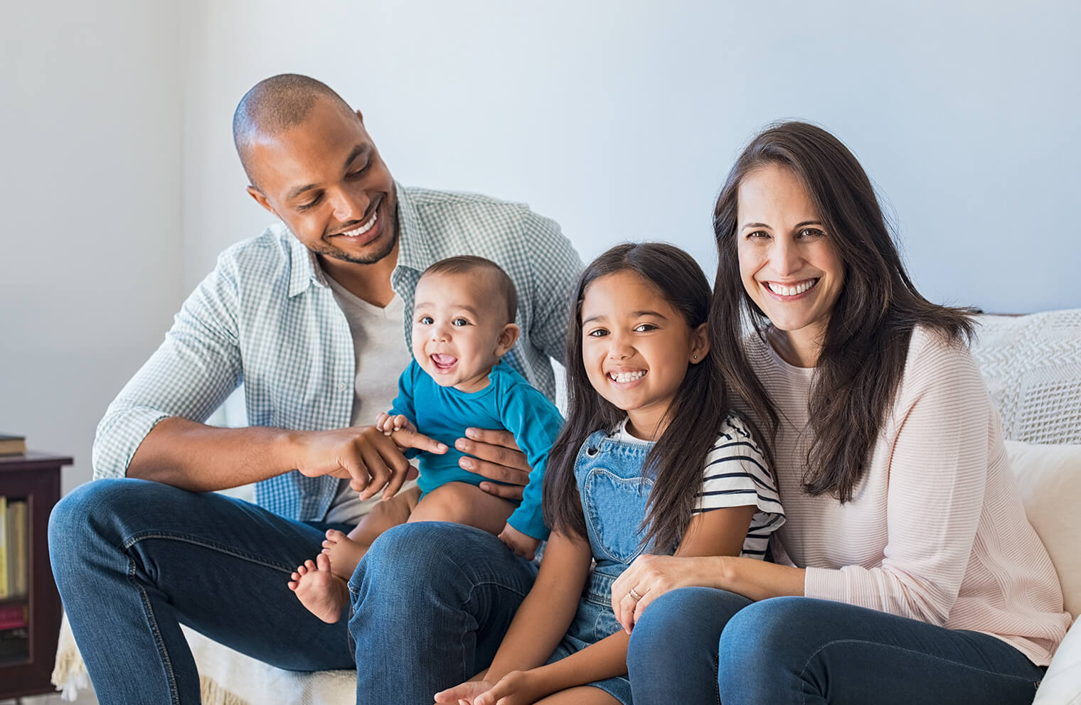 Picture of young, multi-ethnic family smiling and sitting on a couch. From left to right: A father, son, daughter and mother.