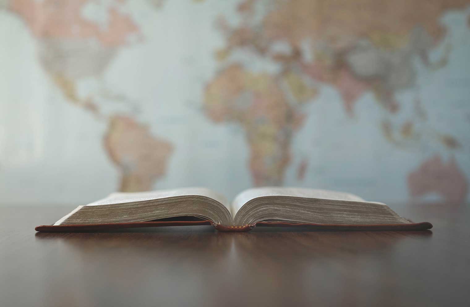An open Bible on a table in front of a wall-sized map of the world