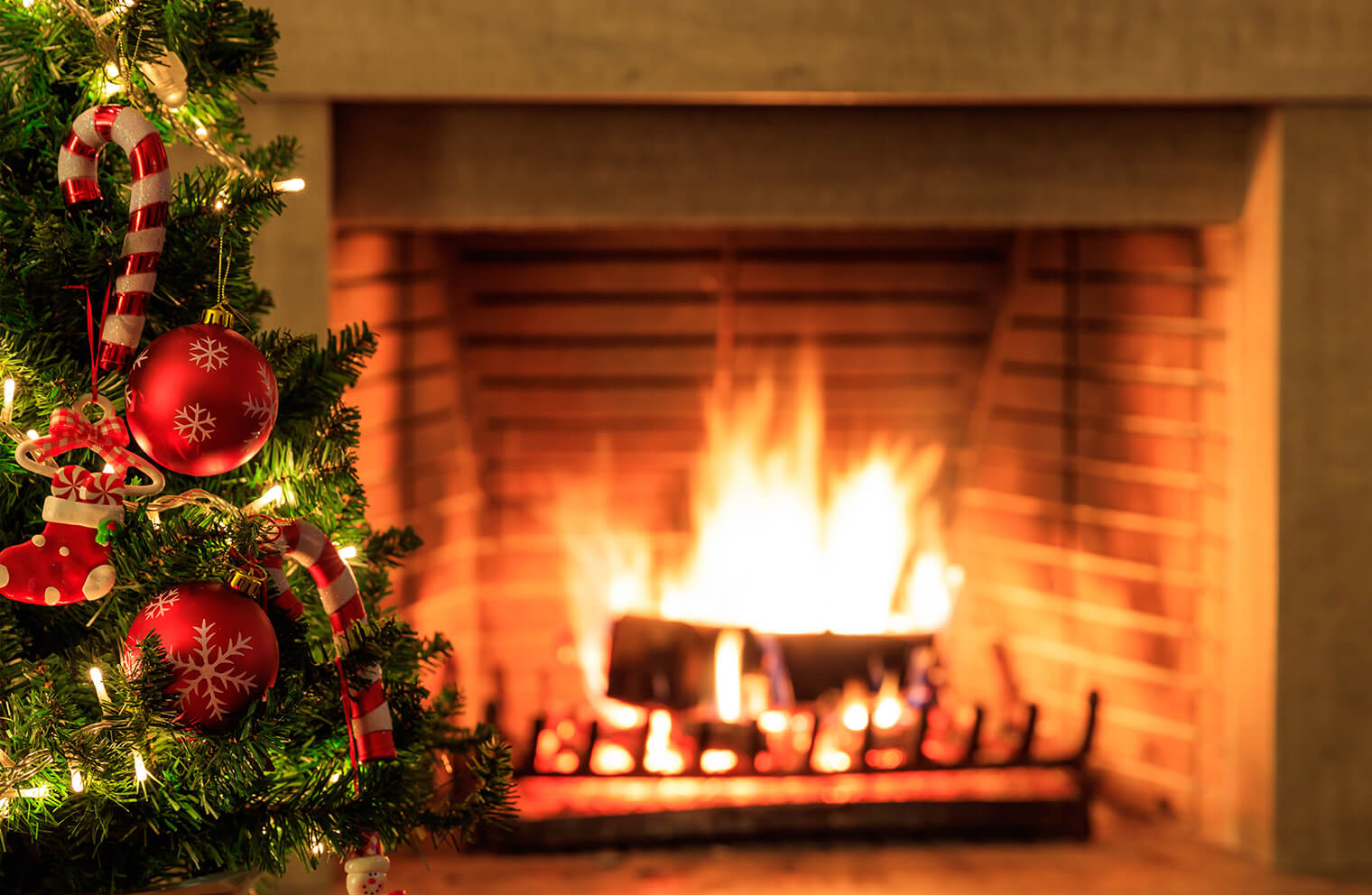 Picture of a Christmas tree that is decorated with red ornaments and candy cane ornaments with a roaring fire in a fireplace in the background.