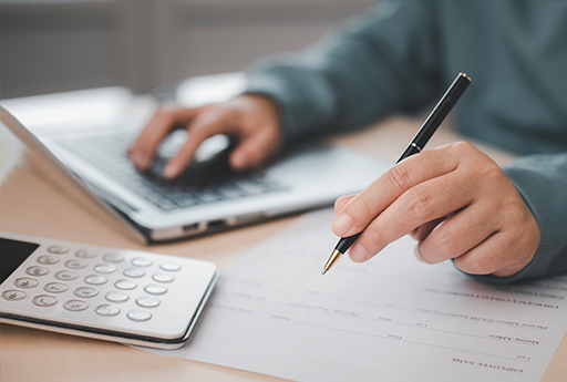 Person writing on a document next to a calculator and laptop keyboard on a desk.