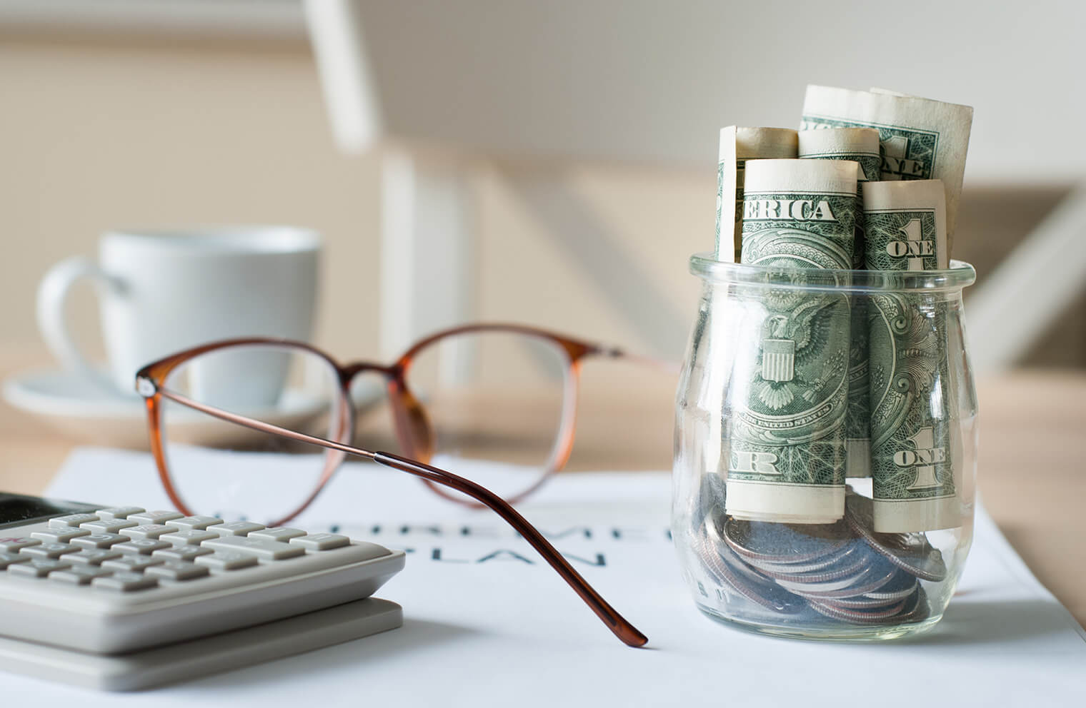 From left to right, a picture of a calculator, pair of glasses, and a small glass jar with coins and bills in it, all on top of a document that says, "Retirement Plan" on it.