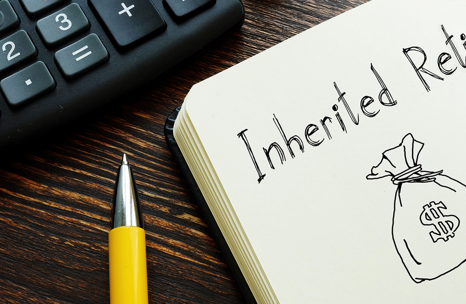 Close up image of the lower third of a calculator, the tip of a pen, and the top, left-hand corner of a journal that has a sketch of a money bag and the beginning of the phrase, "Inherited Retirement".