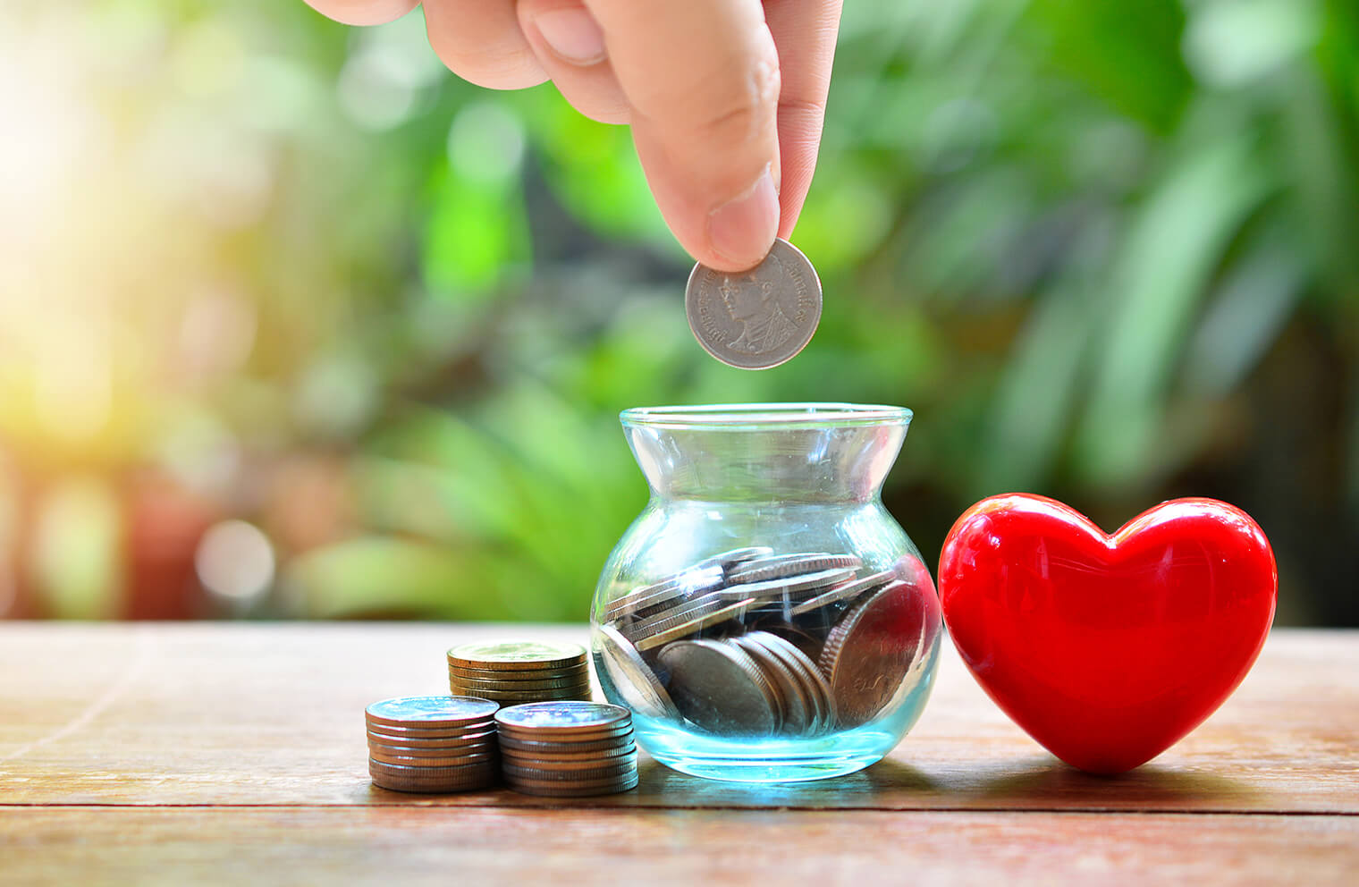 Picture of a person's hand dropping quarters into a tiny glass vase that is next to more quarters to the left and a toy heart to the right.