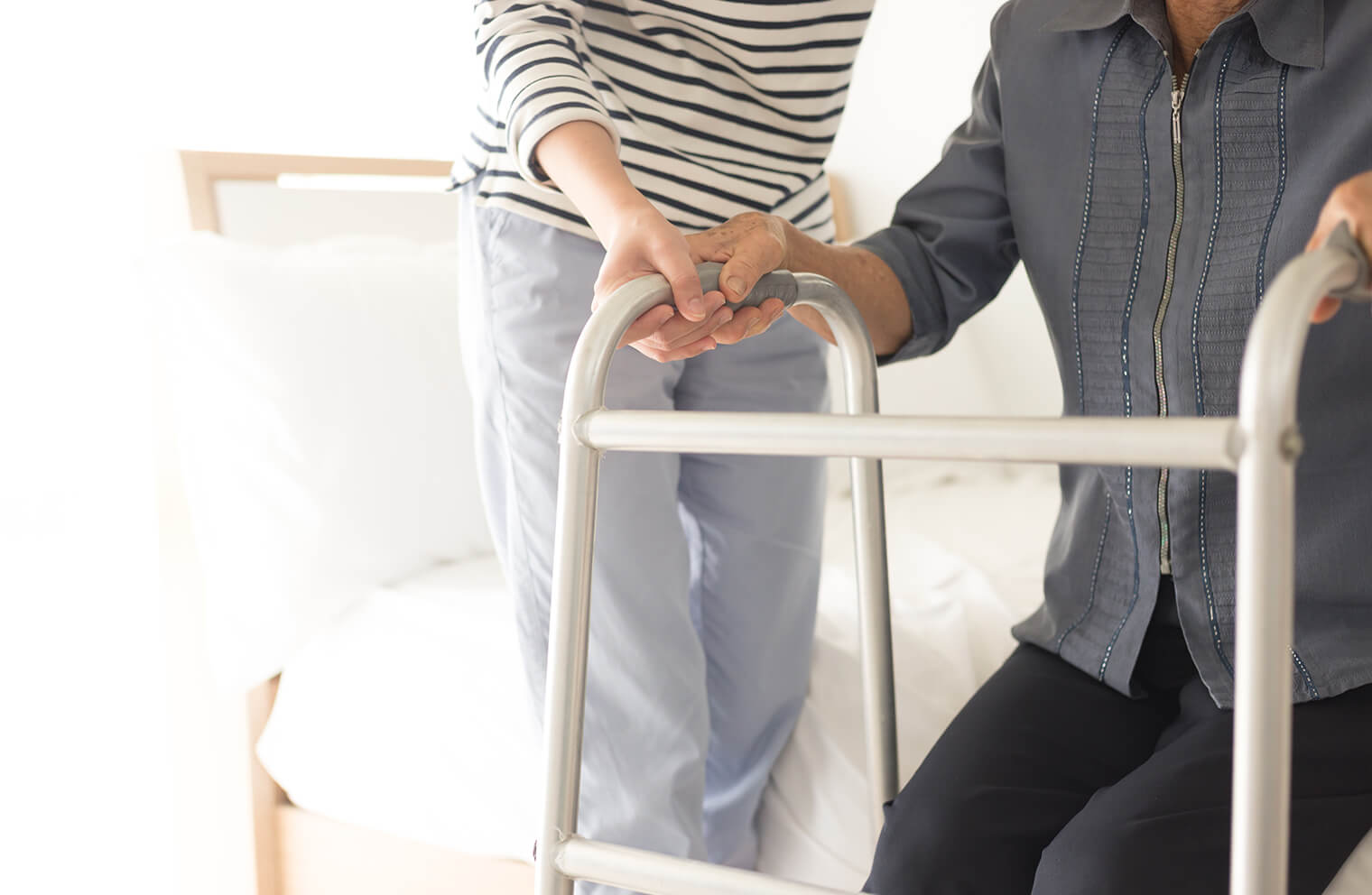 Picture from the neck down of an elderly woman in a navy blouse sitting upright on the edge of a white bed who has her hands on a walker as if she is going to attempt to use it to stand up. There is another woman in a black and white striped blouse and light blue pants on the left, there to help and steady the elderly woman.