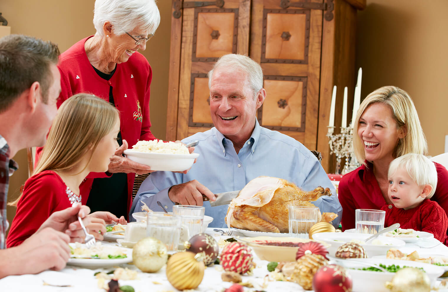 Picture of a family sitting down for Christmas dinner. Man in blue shirt is carving a turkey.