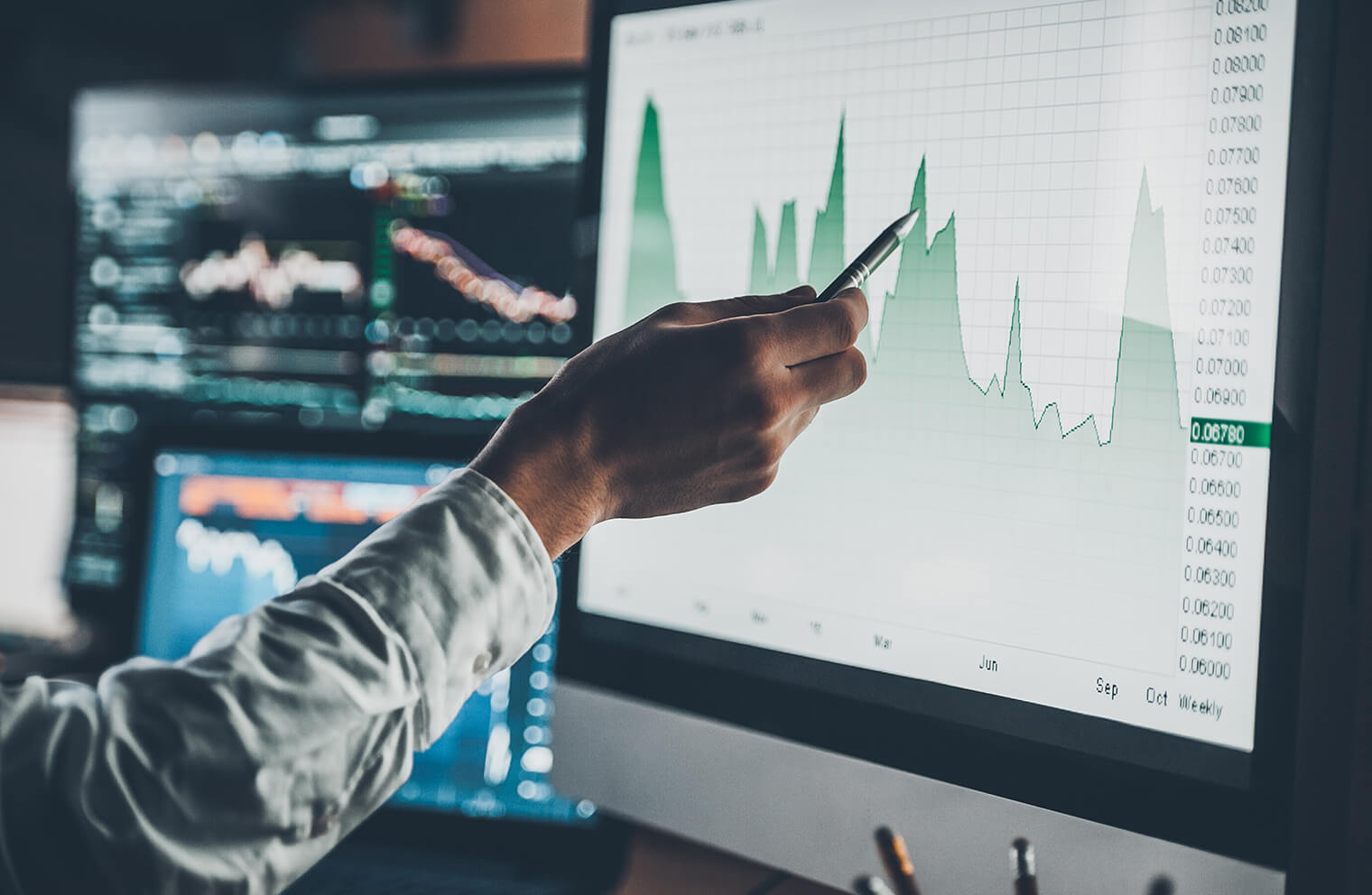 Close up picture of a hand holding a pen and pointing at a line area chart on a computer screen with other computer monitors in the background displaying financial market graphs.
