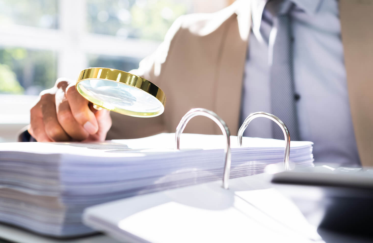 Close up picture of a man in a tan suit with a blue shirt holding a magnifying glass up to a binder of documents.
