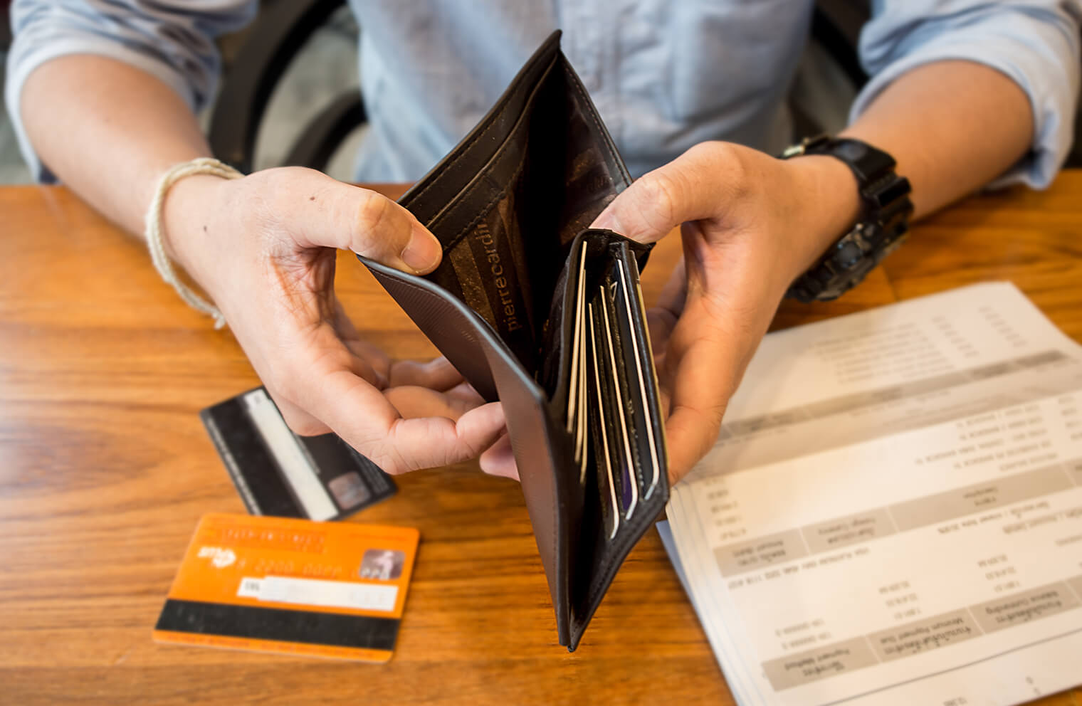 Close up picture of a man holding open an empty wallet on a desk next to a bill.