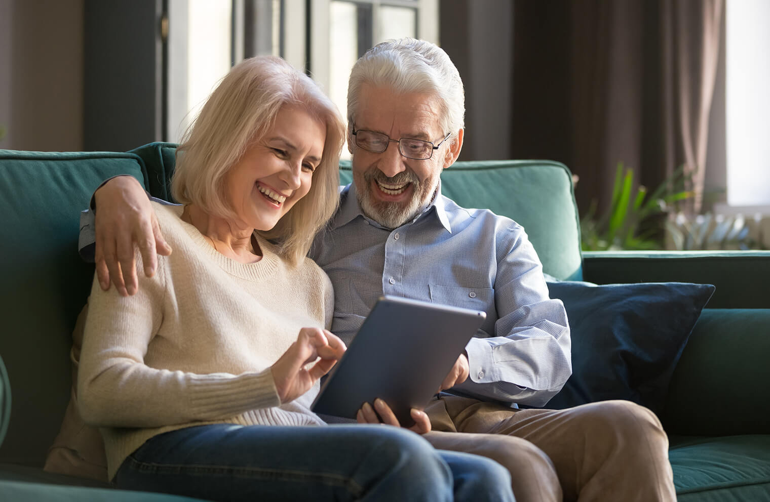 Picture of an older couple sitting on a sage green couch. The husband has his arm around his wife and they are both smiling while looking at a tablet that the wife is holding.