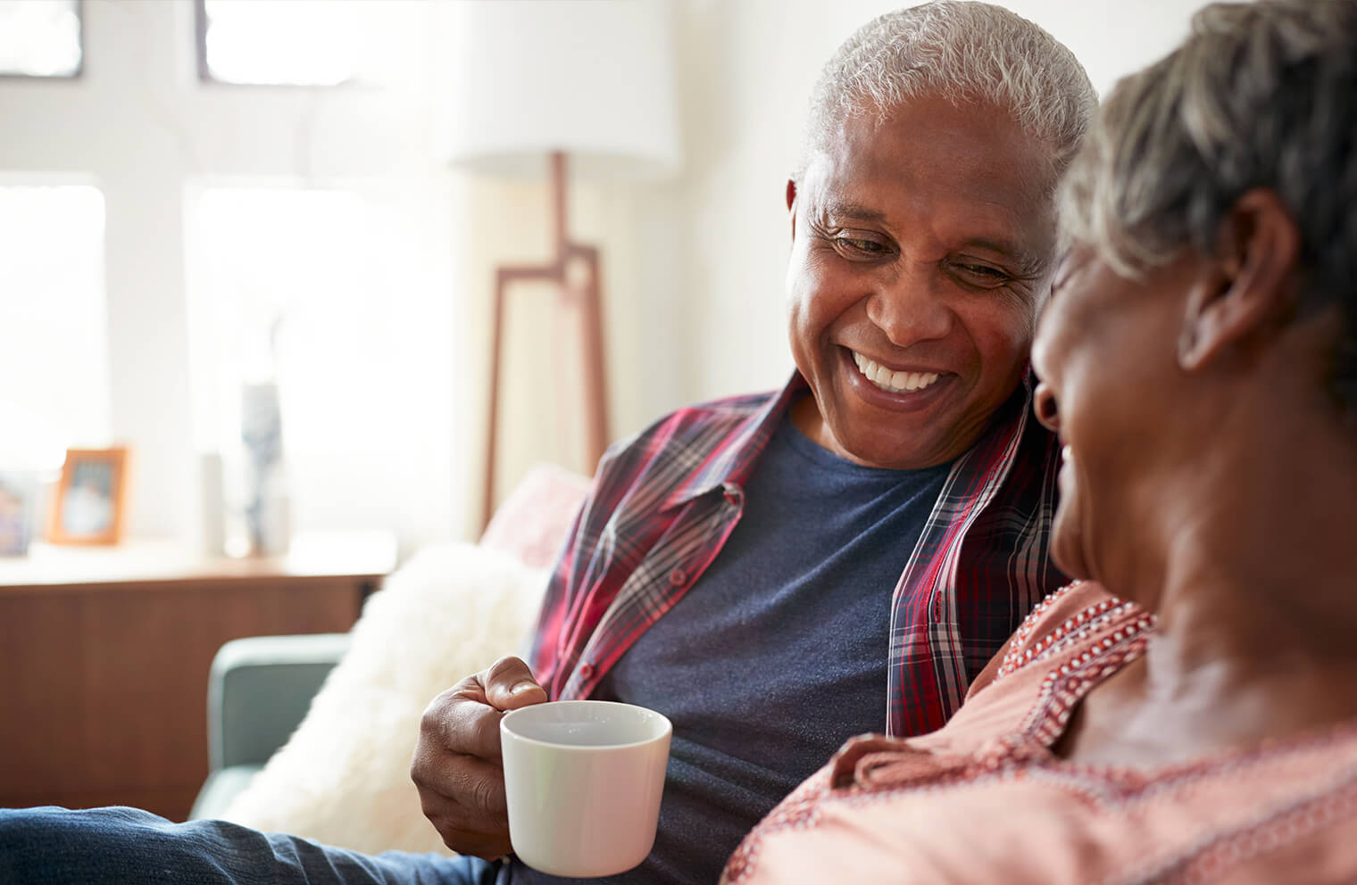 Picture of a retired couple smiling at each other while sitting on a couch and drinking coffee.