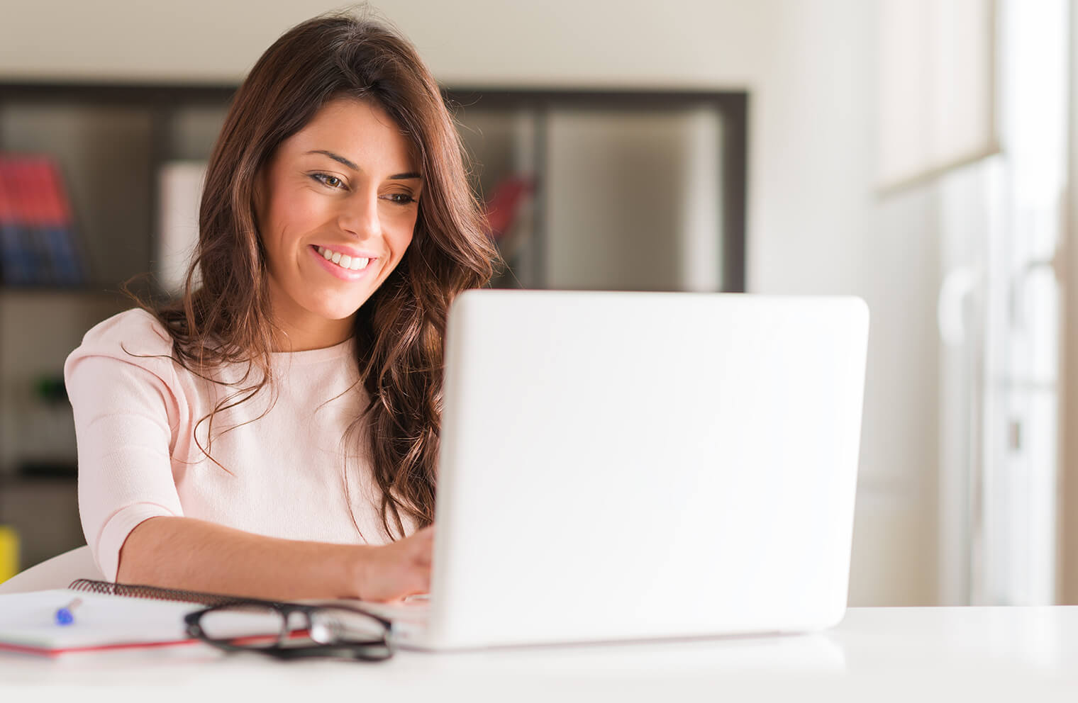 Picture of a woman in a pink shirt smiling while using a laptop on a white countertop with a pair of glasses and pen and paper at her side.