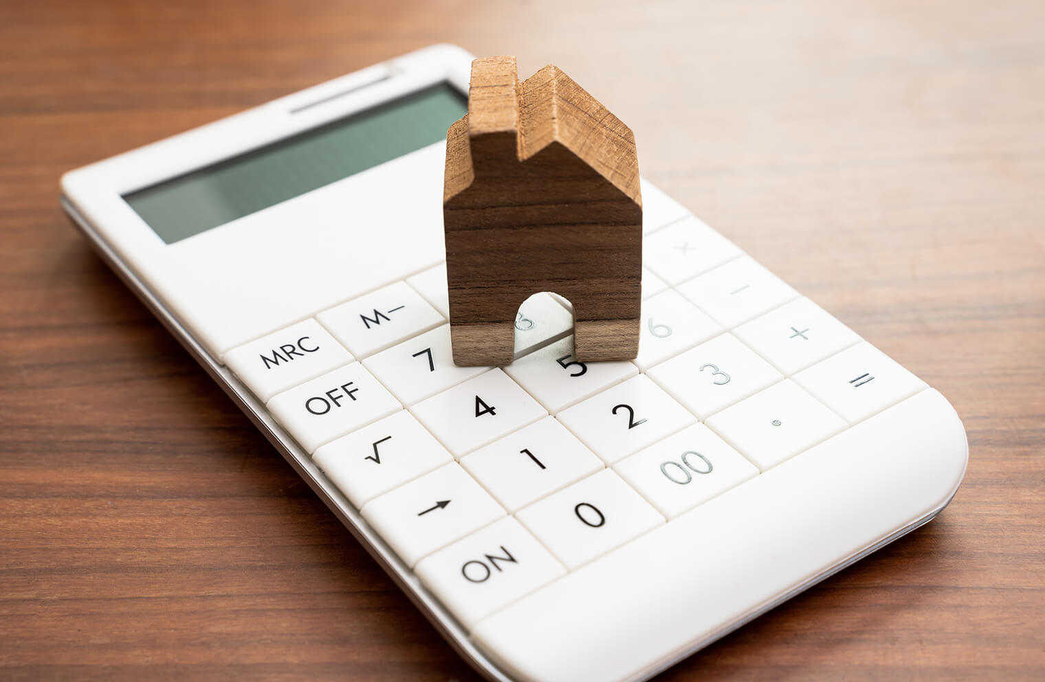 Close up image of a tiny wooden house on top of a white calculator that is on top of a wooden desk.