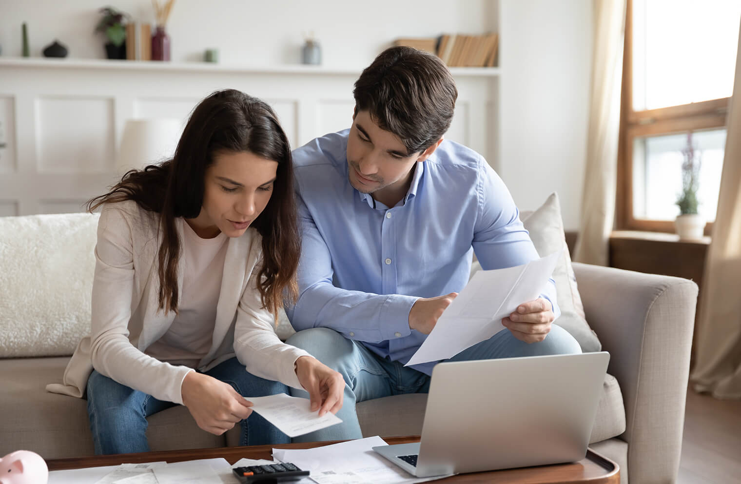 Picture of a young couple sitting on a couch while reviewing bills and receipts. There is a laptop, documents and a calculator on the desk.