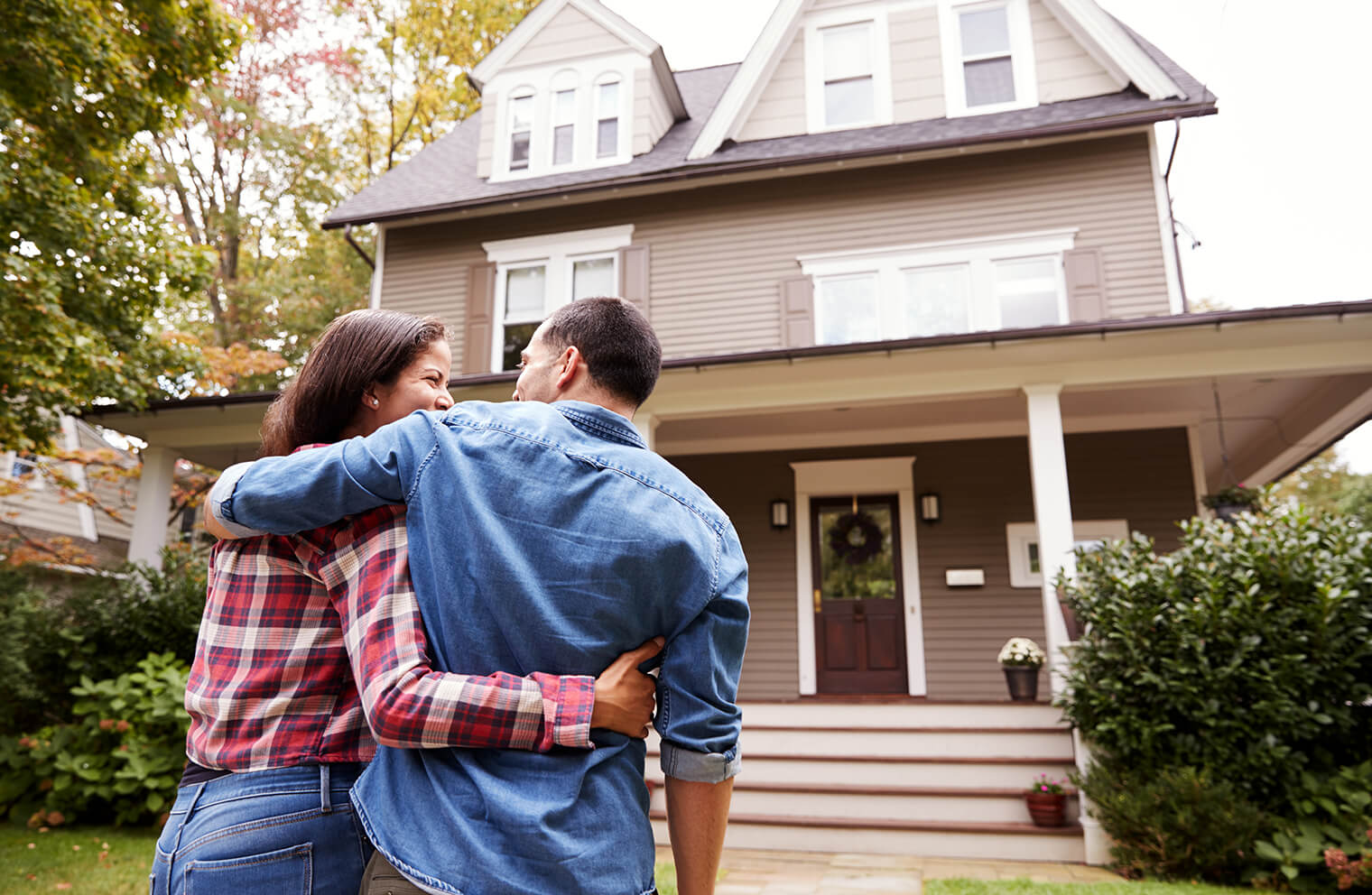 Image from the perspective of being behind a couple who are arm in arm, smiling and looking at each other as they walk toward a three story house.