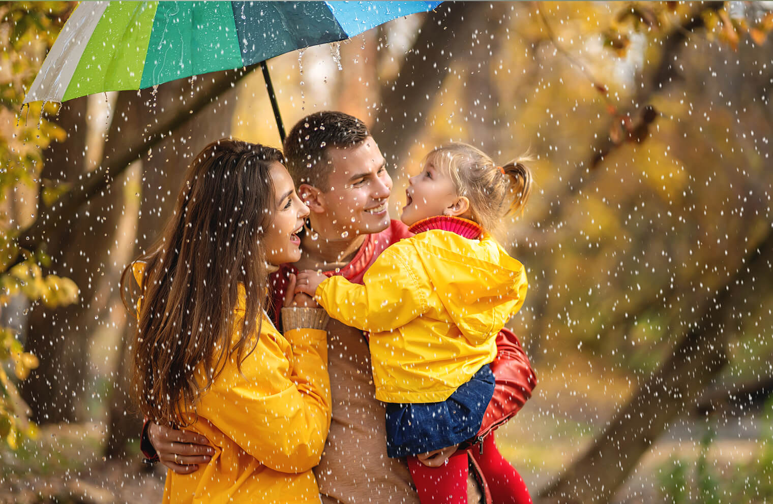 Picture of a young family. Husband, wife and young daughter under an umbrella while it is raining and sunny outside.