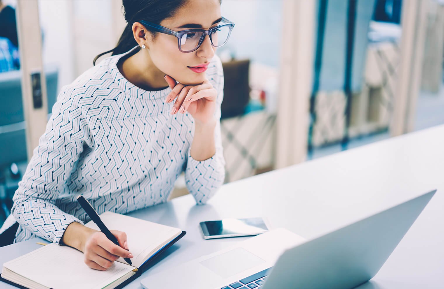 Picture of a young woman in a geometrically patterned white blouse resting her head on her hand while she looks at her laptop on a desk while also taking notes in a journal.